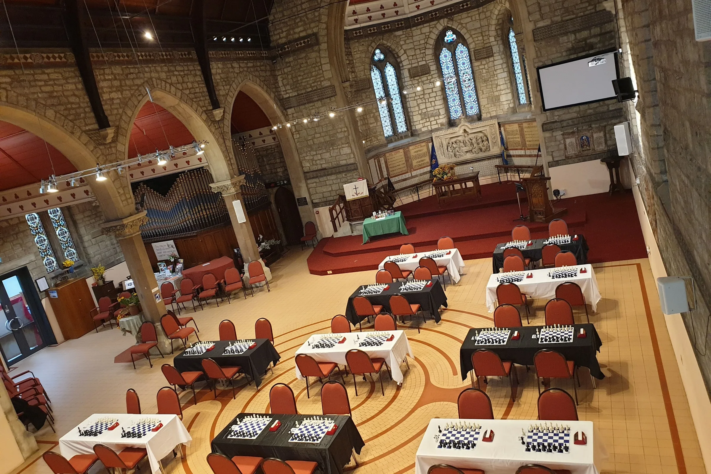 The hall at Chislehurst Methodist Church set up for chess: boards and clocks on tables under stone arches and a stained-glass window.