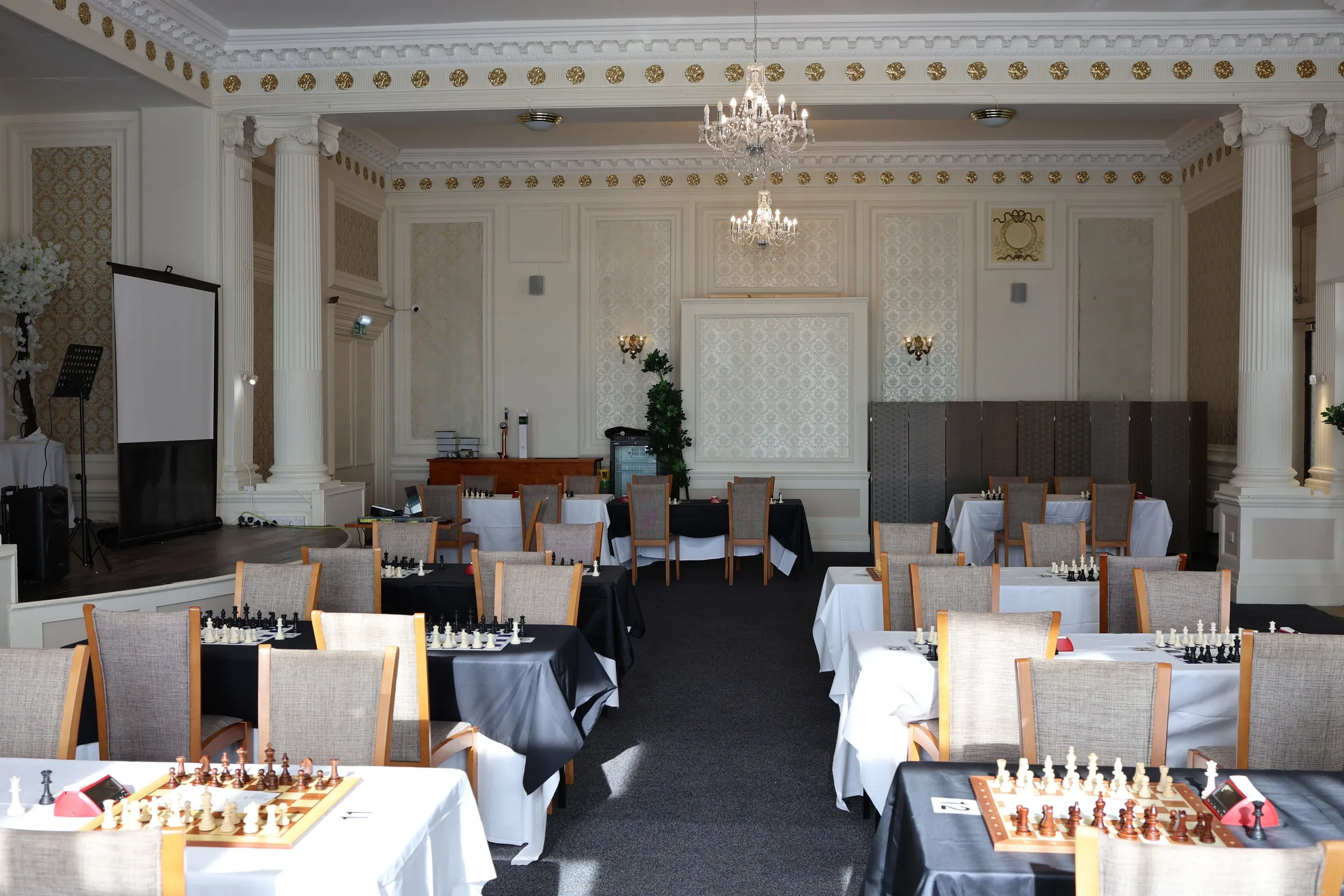 The playing hall at the Grand Burstin Hotel, Folkestone, set up before play: boards and clocks on white- and black-clothed tables beneath chandeliers in a high-ceilinged function room.