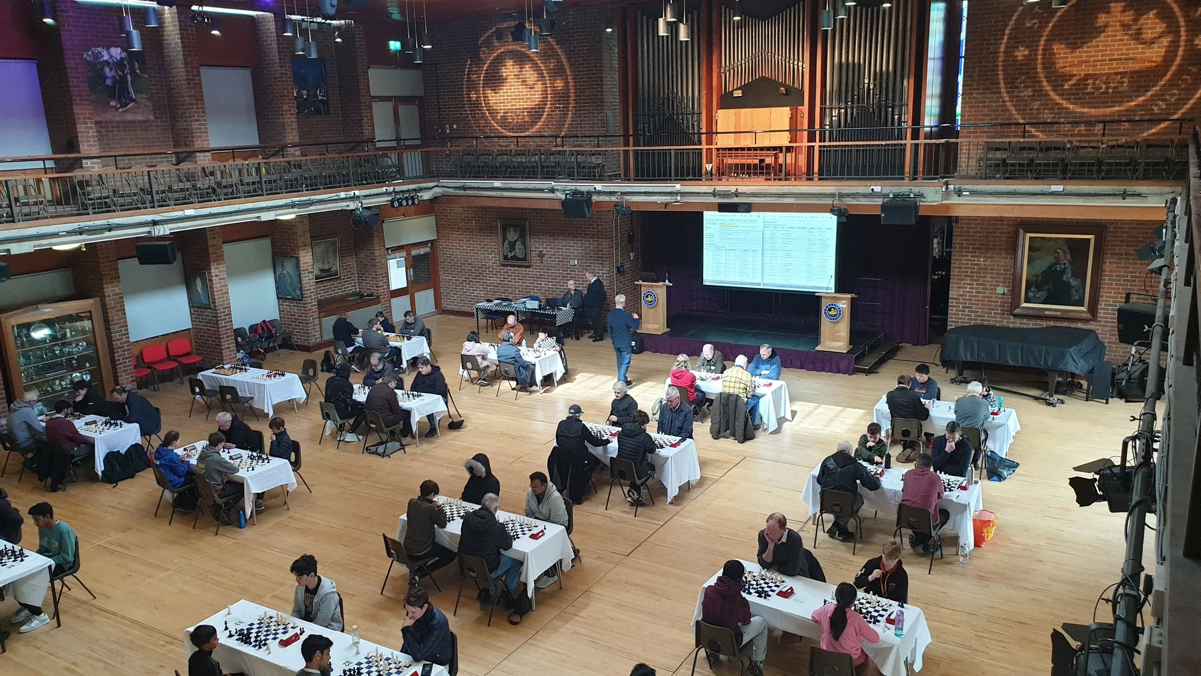 A Psyon Chess congress in play at St Olave's, Orpington: adults and children at white-clothed tables across a large wooden-floored hall, with the live standings shown on a screen above the stage.