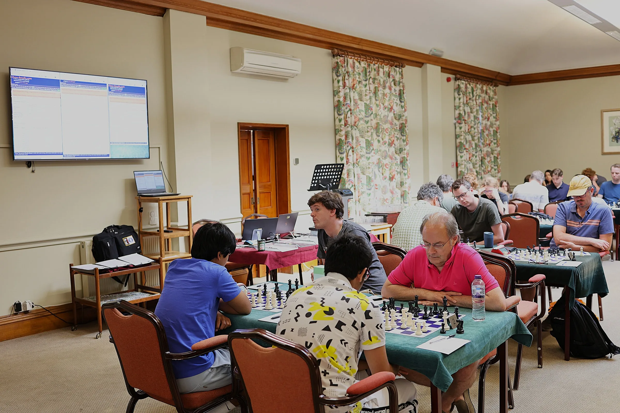 A congress playing hall in use, with players at green baize tables and the live standings on a screen at the far end.